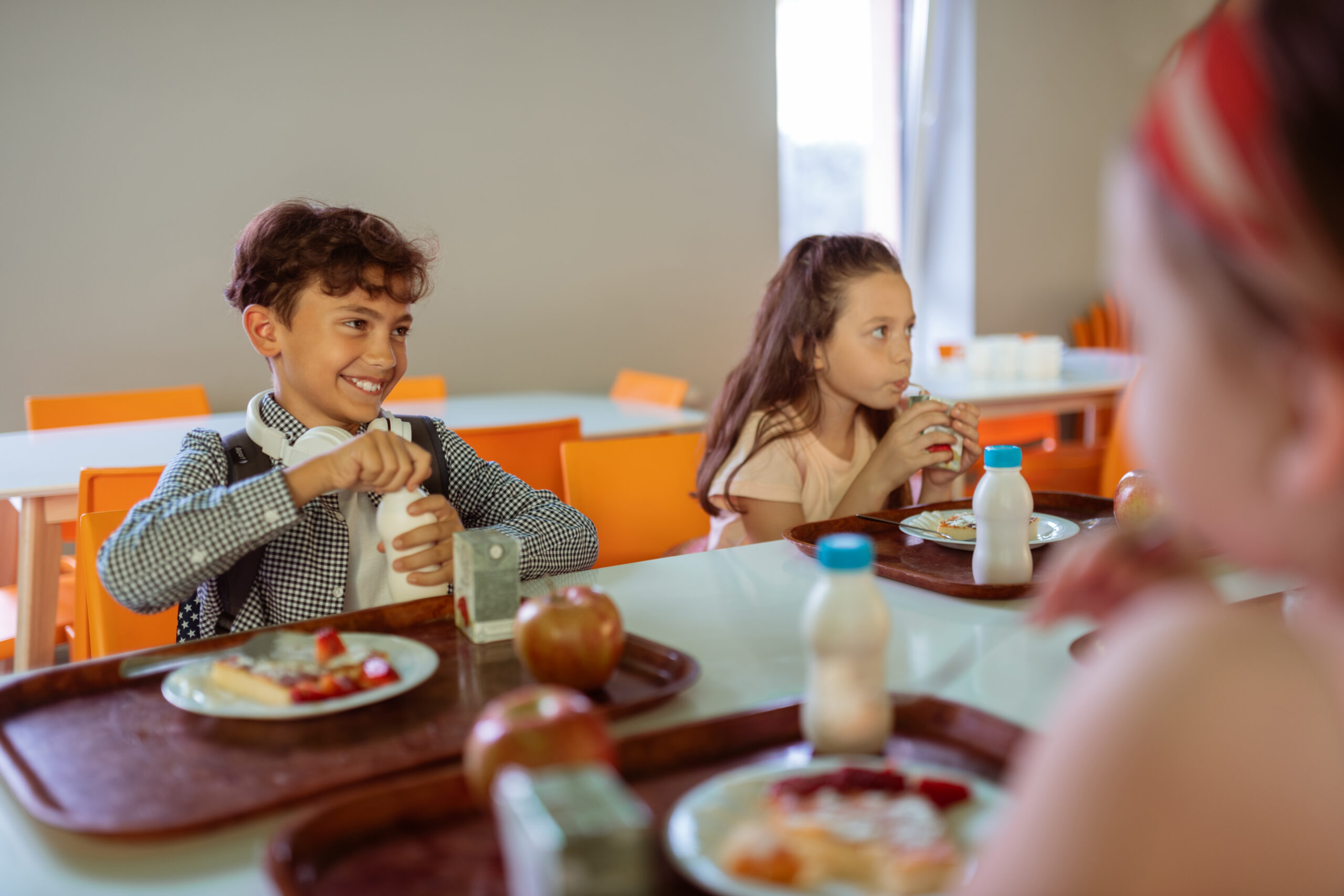 Cheerful boy opening his bottle of milk while having lunch with classmates