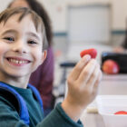 Smiling elementary schoolboy eats strawberries during lunch in the school cafeteria. He is looking at the camera.