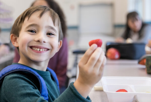 Smiling elementary schoolboy eats strawberries during lunch in the school cafeteria. He is looking at the camera.