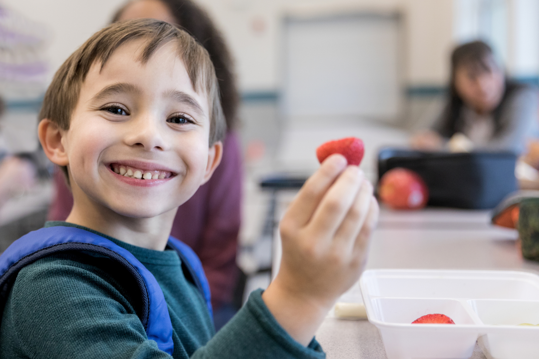 Smiling elementary schoolboy eats strawberries during lunch in the school cafeteria. He is looking at the camera.