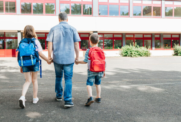 A dad leads his children by the hand with backpacks on their backs to school.