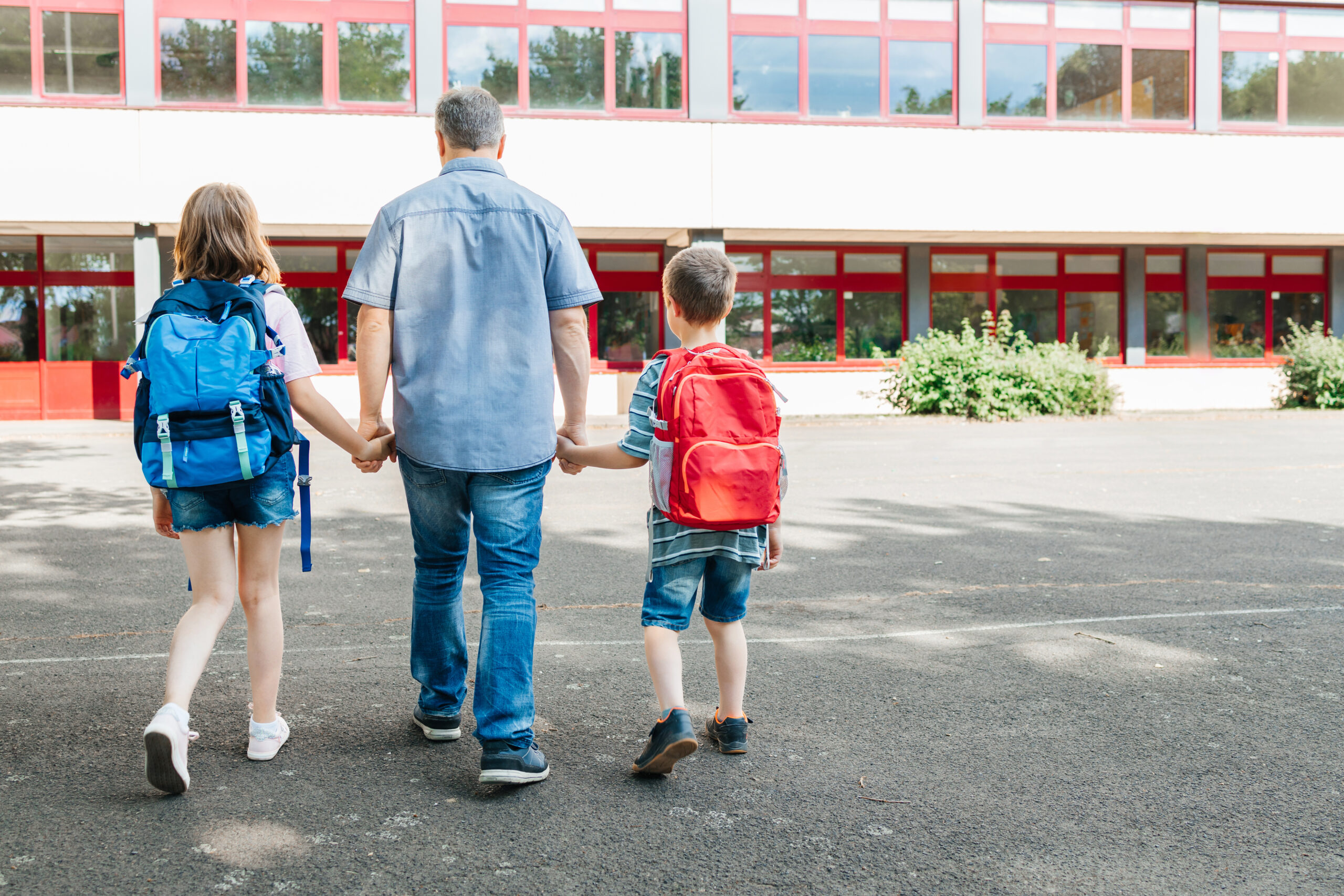 A dad leads his children by the hand with backpacks on their backs to school.
